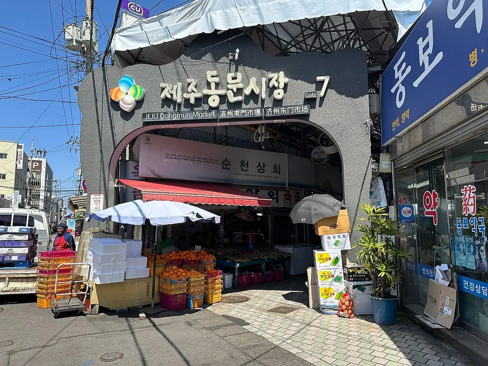 여러 언어의 안내 표지와 과일 가판대가 있는 제주 동문시장 7번 입구